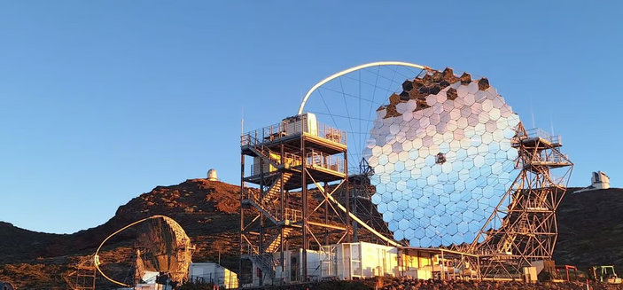 A telescope made of hexagonal mirrors and an oval frame with tensioning wires around it on top of a skeletal frame