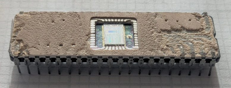 The 8087 chip with the top popped off to reveal the silicon die inside. Complex patterns are visible on the silicon. The chip is a ceramic rectangle. It has 40 metal pins, 20 on each side. The surface of the chip is rough and tan colored.