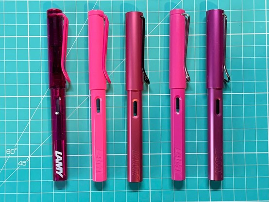 Five fountain pens on a green cutting mat. Each one is a different shade of pink.